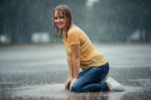 Hannah kneels in a puddle during a heavy rainstorm, smiling at the camera with wet hair and clothes. Her yellow t-shirt, blue jeans and white sneakers are completely soaked.