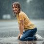 Hannah kneels in a puddle during a heavy rainstorm, smiling at the camera with wet hair and clothes. Her yellow t-shirt, blue jeans and white sneakers are completely soaked.