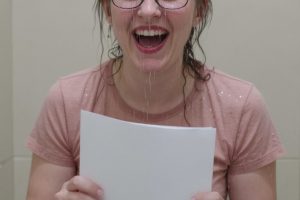 Hannah laughs while sitting on a stool, holding a piece of paper, with water dripping from her hair. She wears a pink t-shirt and grey leggings which are soaked.