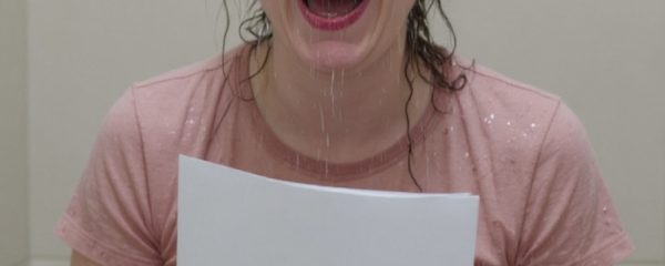 Hannah laughs while sitting on a stool, holding a piece of paper, with water dripping from her hair. She wears a pink t-shirt and grey leggings which are soaked.